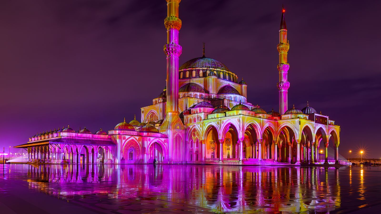 Mosquée illuminée de couleurs vives la nuit, reflétée sur le sol mouillé.