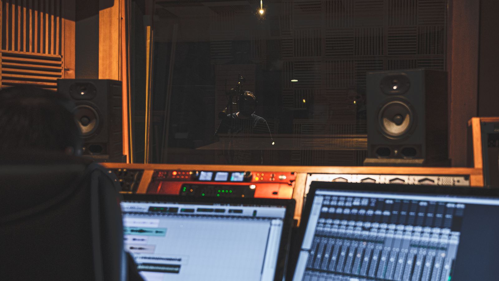 Régie d'un studio d'enregistrement, ingénieur du son de dos face aux écrans, chanteur en cabine derrière la vitre.
