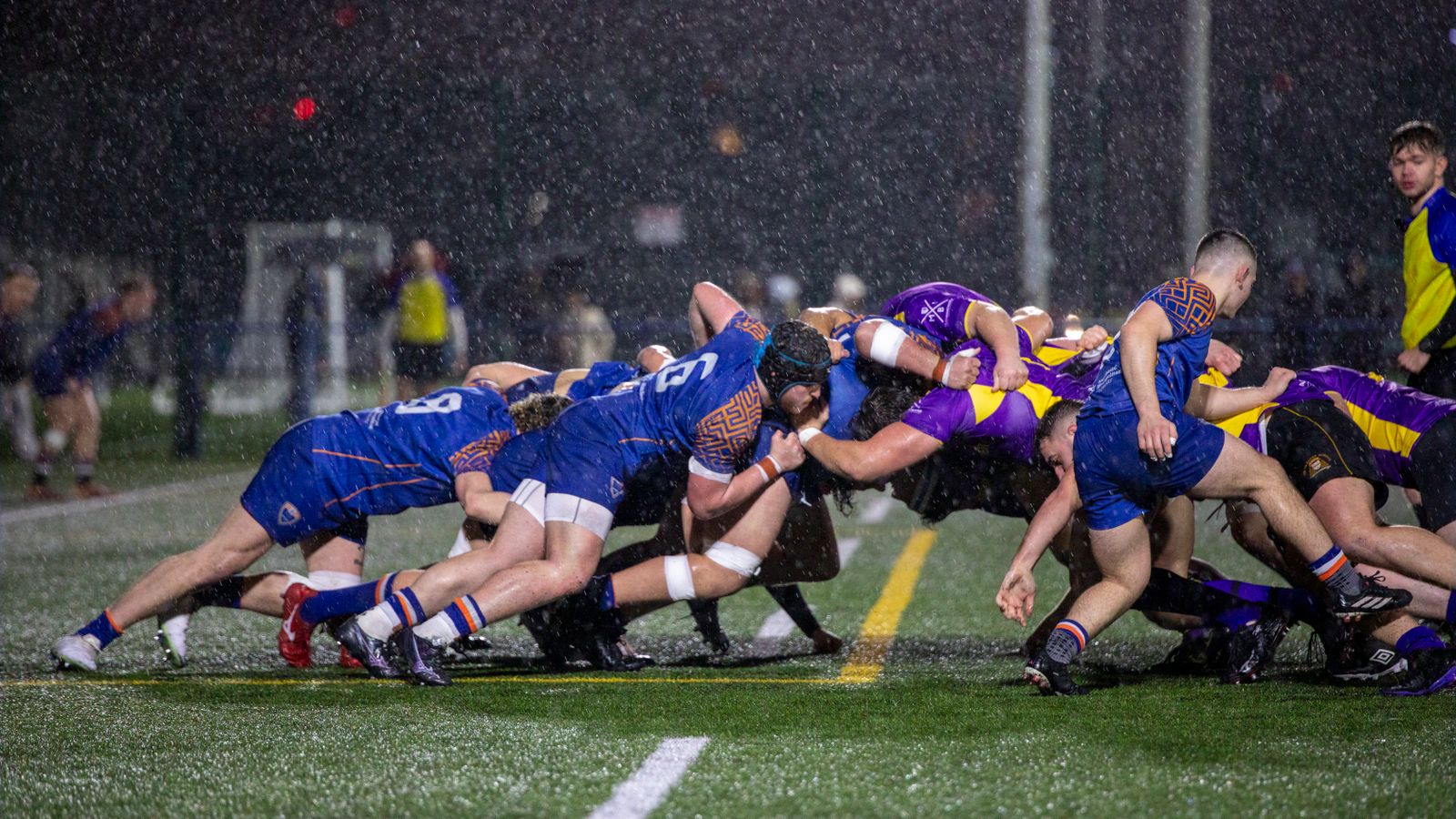 Une mêlée de rugby amateur disputée sous la pluie, de nuit, sur un terrain synthétique.