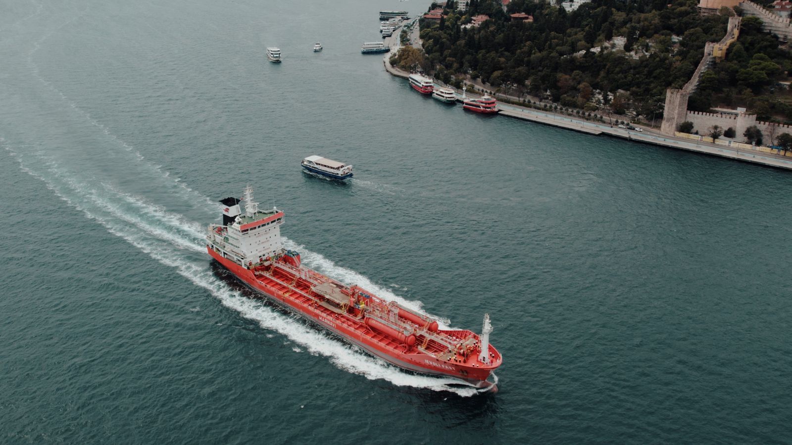Vue aérienne d'un pétrolier naviguant dans un détroit, sillage blanc sur une eau turquoise, ville et bord de mer en arrière-plan.