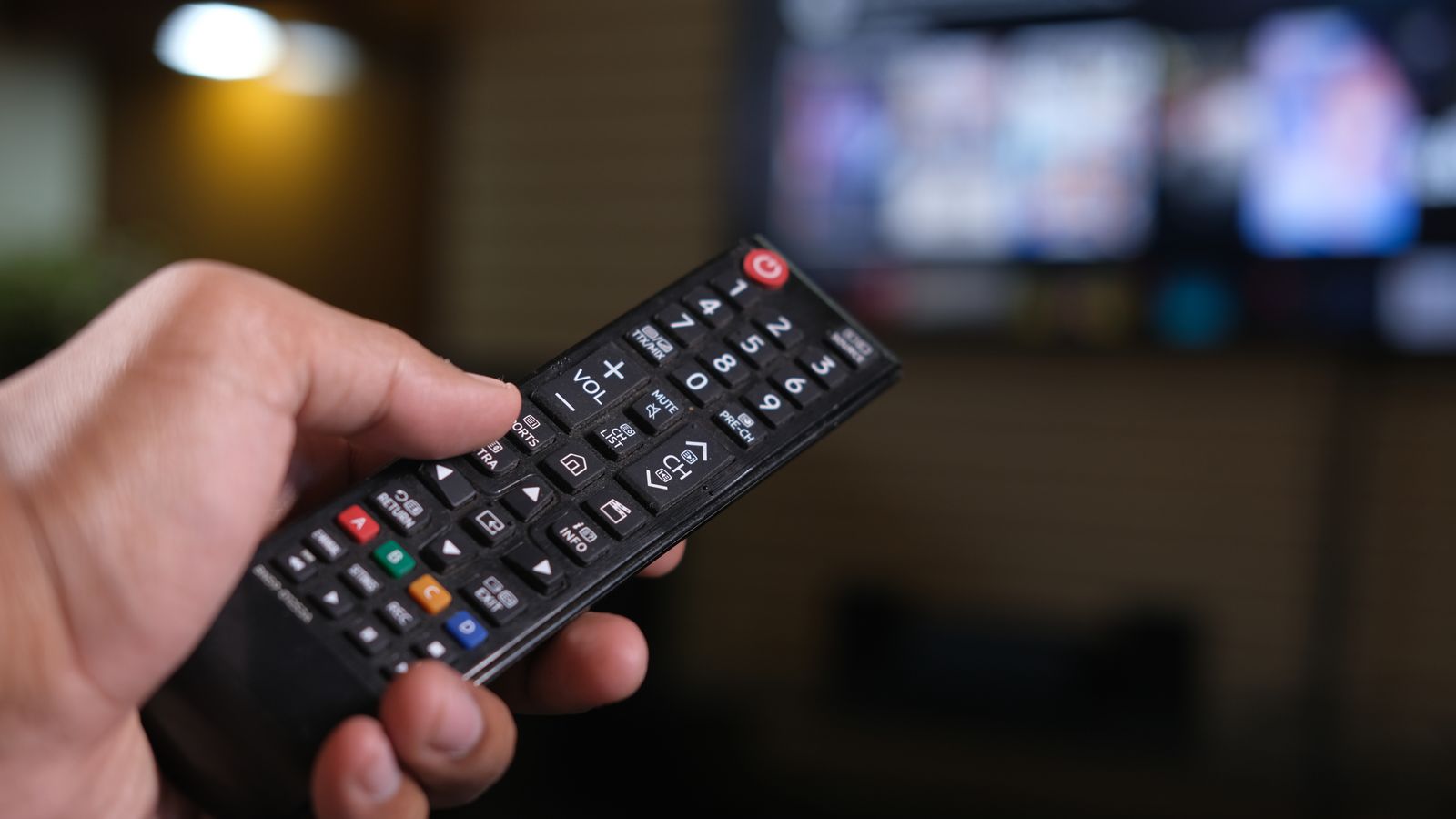 Une main actionne une télécommande devant un écran flouté, dans un salon en soirée.