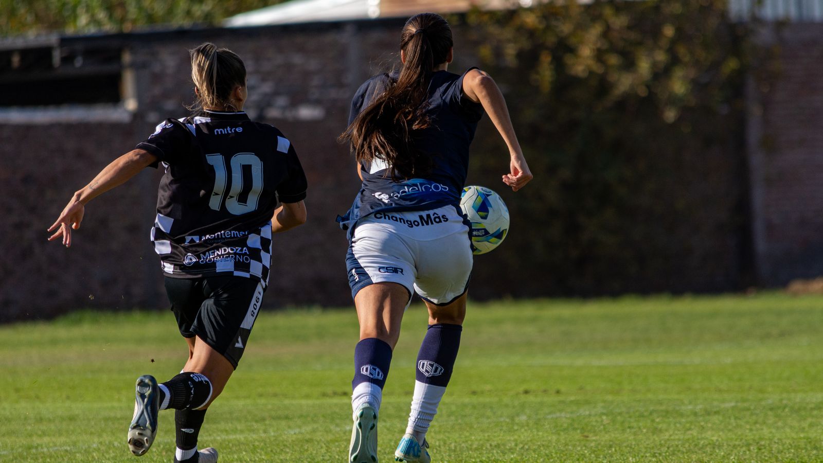 Deux joueuses de football se disputent le ballon sur un terrain en herbe, vues de dos.