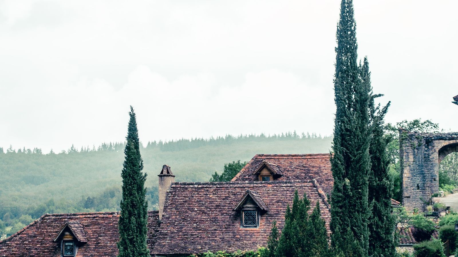 Les toits de tuiles d'un village médiéval français, entouré de collines boisées.